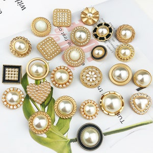 May include: Assortment of gold-toned pearl buttons with various designs, including round, square, heart, and flower shapes. Some buttons have rhinestones or a black trim.
