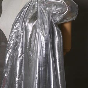 Puede incluir: Una tela plateada y brillante drapeada sobre un maniquí. La tela tiene una apariencia metálica reflectante, con pliegues y arrugas que crean sombras y reflejos. El material probablemente se utiliza para moda o diseño de vestuario.