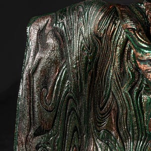 Puede incluir: Tela iridiscente verde y cobre con un patrón de mármol en espiral. La tela tiene un brillo metálico y una superficie texturizada.