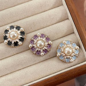 May include: Three decorative round brooches with pearl and rhinestone accents. One brooch is black and white, another is purple and white, and the third is light blue and white. Each brooch has a large central pearl.