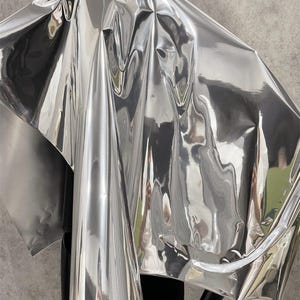Peut inclure: Un tissu argenté brillant et réfléchissant drapé sur un mannequin noir. Le tissu présente des plis et des reflets, créant un effet de texture. L'ensemble dégage une esthétique moderne et abstraite.