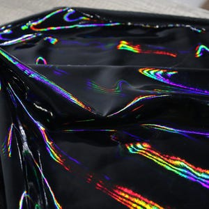 Peut inclure: Un morceau de tissu noir avec une finition brillante et réfléchissante. Le matériau présente un effet holographique qui affiche des traînées de lumière multicolores vibrantes dans des tons de vert, jaune et violet sur sa surface lisse et pliée.