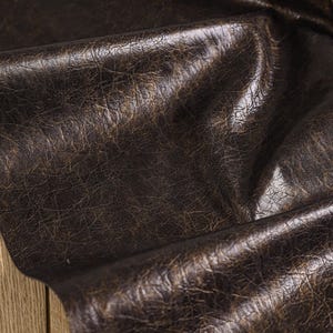 May include: Close-up of a dark brown leather material with a distressed, textured appearance. The leather has a rich, aged look with subtle variations in colour and a slight sheen. The folds and creases add depth.