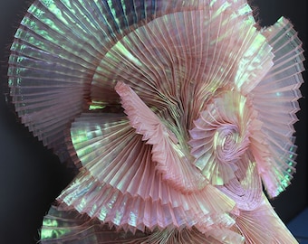 May include: A large, pink, iridescent, pleated fabric sculpture. The fabric is folded and layered to create a three-dimensional, organic shape.