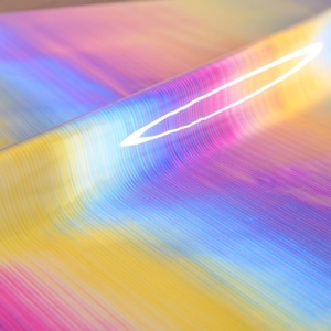 May include: A sheet of glossy material featuring a vibrant, multi-colored gradient pattern with horizontal lines. The colors transition smoothly from deep purple and magenta to bright yellow and light blue, creating a reflective, iridescent surface.