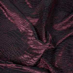 May include: A close-up of a deep burgundy fabric with a crocodile skin texture. The fabric has a sheen, with highlights and shadows that create depth. The material is folded, showing the texture and color variations.