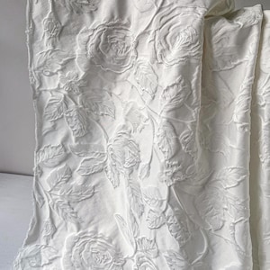 May include: White fabric with a raised floral pattern of roses and leaves. The fabric is folded, showing the texture and design. The roses are in various stages of bloom, creating a detailed and elegant look.