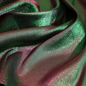 May include: Close-up of shimmering fabric with a blend of green and reddish-purple hues. The material is folded, creating shadows and highlights that emphasize its texture and sheen. The fabric appears to be soft and flowing.