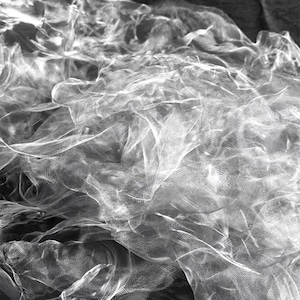May include: A monochrome image showcasing a mass of sheer, flowing fabric. The material is layered and textured, creating a sense of volume. The light filters through the delicate weave, highlighting the intricate details of the fabric.