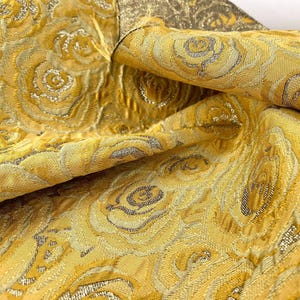 Puede incluir: Primer plano de una tela amarillo dorado con un patrón de rosas repetido. La tela tiene un brillo metálico y una superficie texturizada. Las rosas están delineadas en plata, agregando contraste al diseño. La tela está doblada, revelando el reverso.