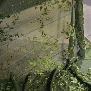 May include: Dark green sheer fabric with an embroidered floral pattern. The design features detailed leaves and vines in varying shades of green, creating a textured, botanical aesthetic. The fabric is draped, showing its delicate, flowing nature.