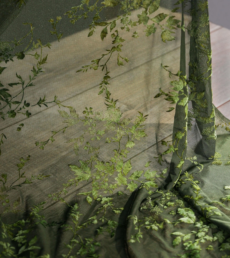 May include: Dark green sheer fabric with an embroidered floral pattern. The design features detailed leaves and vines in varying shades of green, creating a textured, botanical aesthetic. The fabric is draped, showing its delicate, flowing nature.