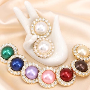 May include: A set of eight decorative pearl and rhinestone buttons in various colours, including green, blue, purple, pink, red, and brown. The buttons are arranged in a row on a white surface.