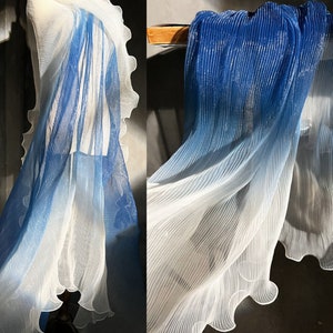 May include: A sheer, pleated fabric in a gradient of blue and white. The fabric has a wavy, ruffled edge and is draped to show its texture and colour transition. The material appears lightweight and flowing.