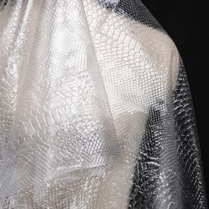May include: A white, translucent fabric with a crocodile skin pattern. The fabric is draped over a form and has a slight sheen.