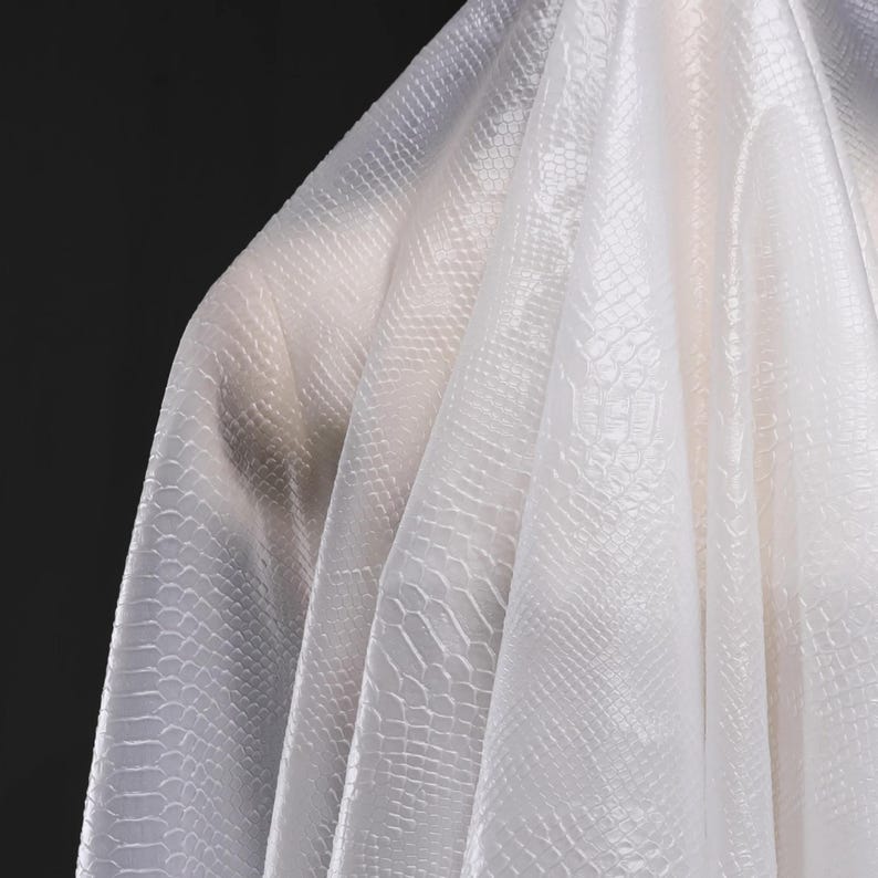 May include: White fabric with a textured, embossed pattern resembling crocodile skin. The fabric is translucent and drapes softly.