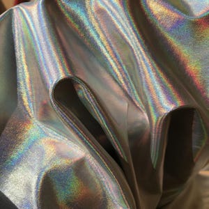 Peut inclure: Un morceau de tissu avec une finition holographique métallique qui reflète un spectre de couleurs arc-en-ciel. Le matériau est drapé pour créer des plis profonds et des ombres, soulignant la surface lisse et réfléchissante ainsi que les motifs lumineux changeants.
