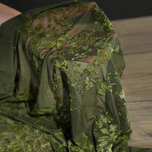 May include: Olive green sheer fabric with a floral pattern. The fabric is held up, showing the delicate texture and design. The floral pattern is a lighter shade of green, creating a contrast against the darker base. This fabric is suitable for apparel and craft projects.
