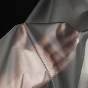 May include: A sheet of translucent, grey fabric is held up, revealing the hand behind it. The fabric has a smooth texture and drapes in soft folds. The background is a dark, solid colour.