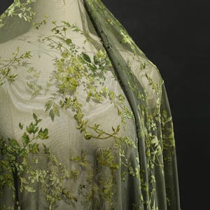 May include: Green sheer fabric with embroidered floral and leaf patterns. The fabric is draped over a mannequin, showcasing the delicate, detailed design. The embroidery features varying shades of green, creating a textured, dimensional effect.