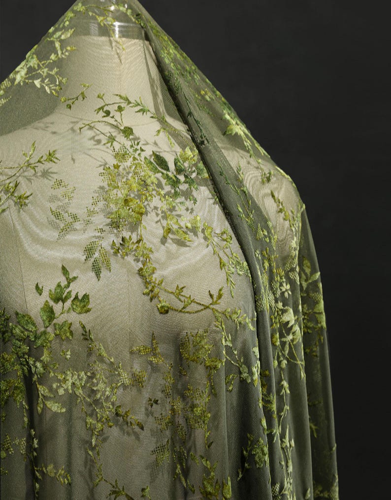 May include: Green sheer fabric with embroidered floral and leaf patterns. The fabric is draped over a mannequin, showcasing the delicate, detailed design. The embroidery features varying shades of green, creating a textured, dimensional effect.