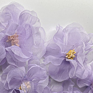 May include: A cluster of delicate, lavender-colored fabric flowers. The flowers are made of sheer, layered fabric and have a slightly ruffled appearance. The flowers are arranged in a close-up view, with the center of each flower visible.