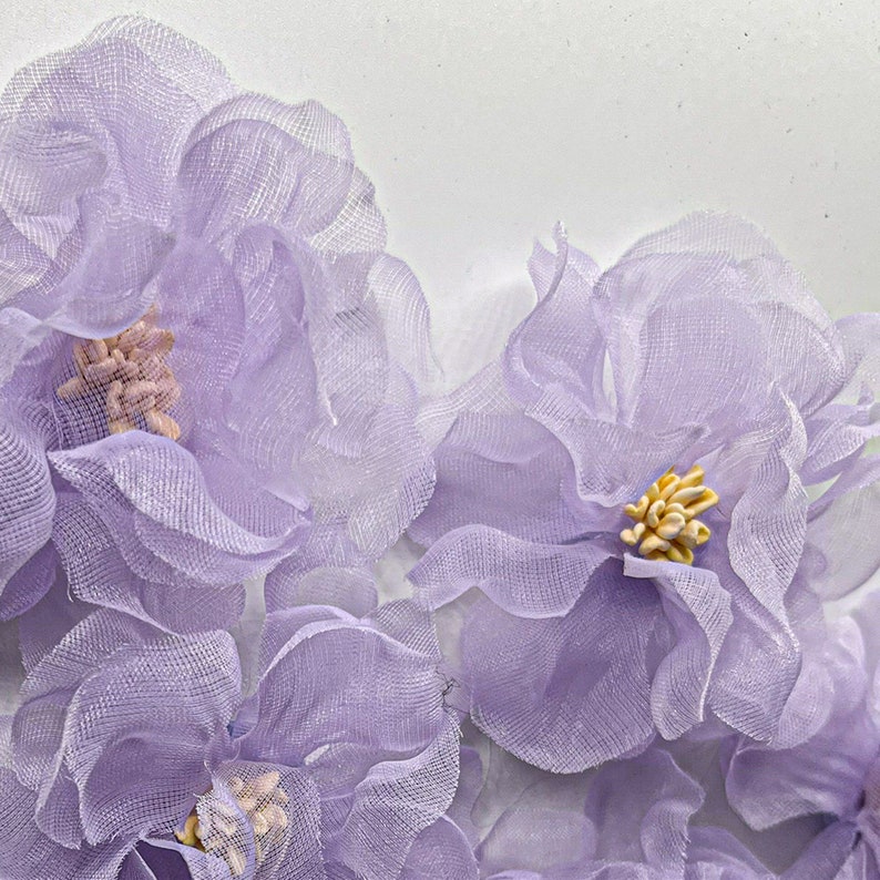 May include: A cluster of delicate, lavender-colored fabric flowers. The flowers are made of sheer, layered fabric and have a slightly ruffled appearance. The flowers are arranged in a close-up view, with the center of each flower visible.