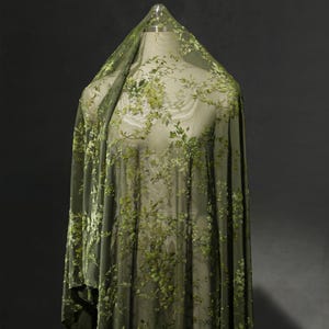 May include: Olive green sheer fabric with embroidered floral patterns. The fabric is draped over a mannequin, showcasing the delicate floral design. The embroidery features varying shades of green, creating a textured, botanical look.