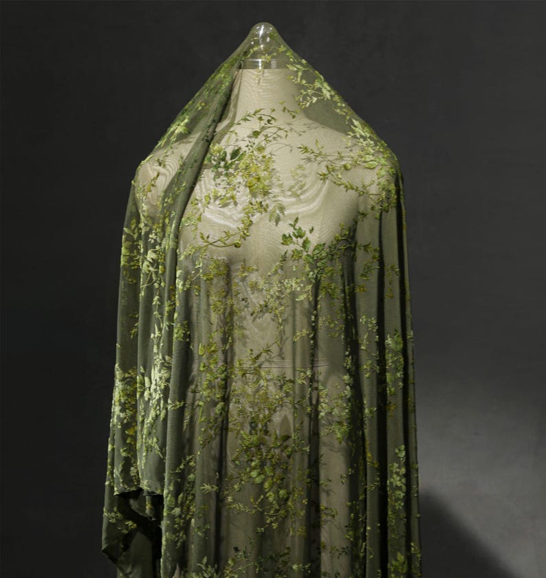 May include: Olive green sheer fabric with embroidered floral patterns. The fabric is draped over a mannequin, showcasing the delicate floral design. The embroidery features varying shades of green, creating a textured, botanical look.