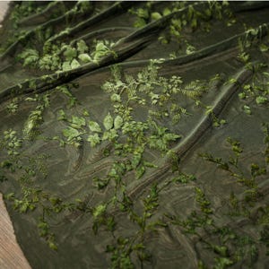 May include: Dark olive green fabric with an intricate floral design. The fabric features raised, three-dimensional leaves and vines in varying shades of green, creating a textured, botanical pattern. The fabric is draped, showing the detailed design.