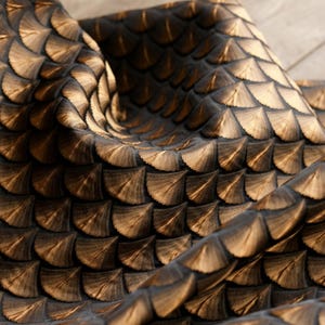 May include: Close-up of a fabric with a repeating pattern of overlapping, fan-shaped elements. The fabric has a dark brown base colour with lighter brown highlights, creating a textured, scale-like appearance. The fabric is draped, showing depth and dimension.