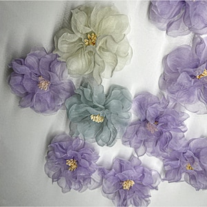 May include: A collection of nine delicate fabric flowers in shades of purple, green, and cream. The flowers are made of sheer fabric and have a layered, ruffled appearance.