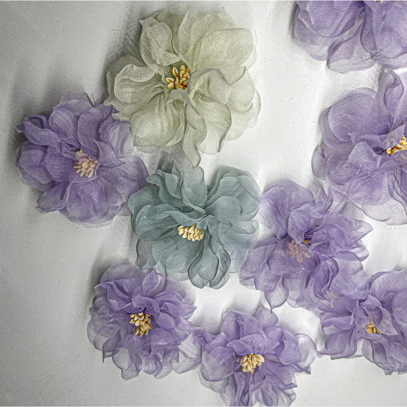 May include: A collection of nine delicate fabric flowers in shades of purple, green, and cream. The flowers are made of sheer fabric and have a layered, ruffled appearance.