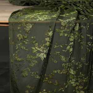 May include: Dark olive green fabric with a floral pattern. The fabric is draped over a wooden surface, showcasing the intricate design of leaves and flowers in varying shades of green. The texture appears soft and delicate, suitable for apparel or home decor.