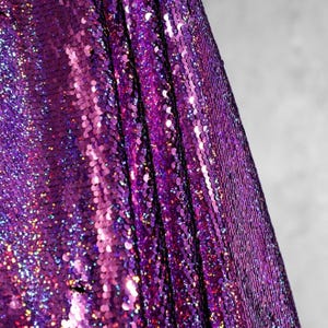 May include: Close-up of a fabric covered in shimmering purple sequins. The sequins are densely packed, creating a textured surface that reflects light. The fabric has a slight drape, with vertical folds visible.