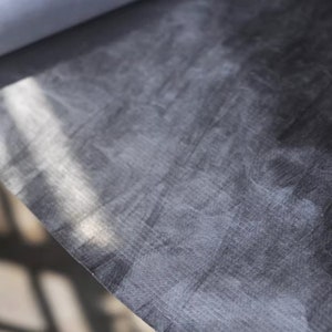 May include: Close-up of a dark gray fabric with a subtle textured pattern. The fabric has a slightly wrinkled appearance and is illuminated by a soft light source.
