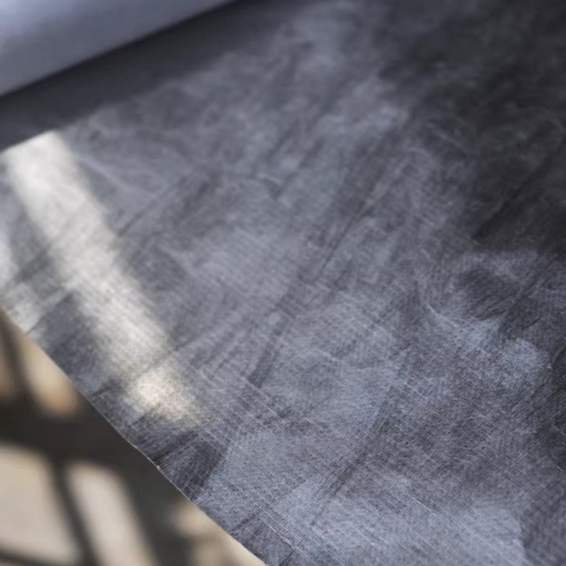May include: Close-up of a dark gray fabric with a subtle textured pattern. The fabric has a slightly wrinkled appearance and is illuminated by a soft light source.