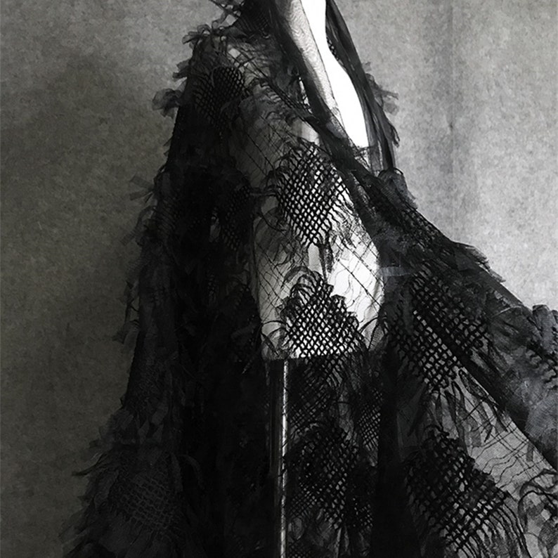 May include: Black lace fabric with a textured, fringed design. The fabric is draped over a mannequin.