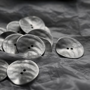 以下が含まれることがあります： しわくちゃの白い背景に置かれた、質感のある波打った表面を持つ銀色のボタンの山をクローズアップした写真です。