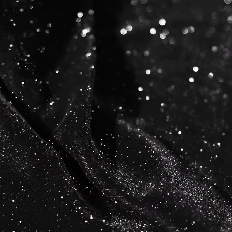 May include: Black fabric with a sparkly, glittery texture. The fabric is draped and folded, creating a soft, flowing look.
