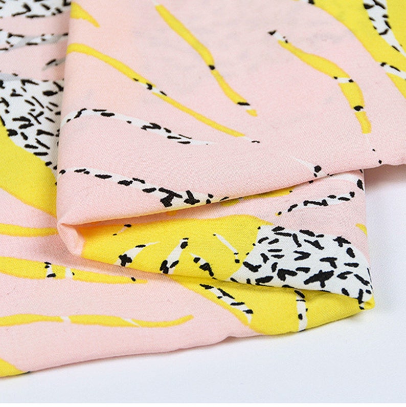 Op de afbeelding: Roze stof met een abstracte print in geel en zwart. De stof is dubbelgevouwen, waardoor het patroon aan beide kanten zichtbaar is.
