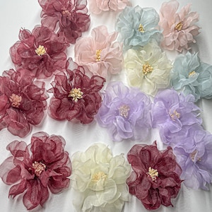 May include: Assortment of 12 delicate fabric flowers in various colors, including burgundy, lavender, cream, and light blue. The flowers are made of a soft, sheer fabric and have a layered, ruffled appearance.