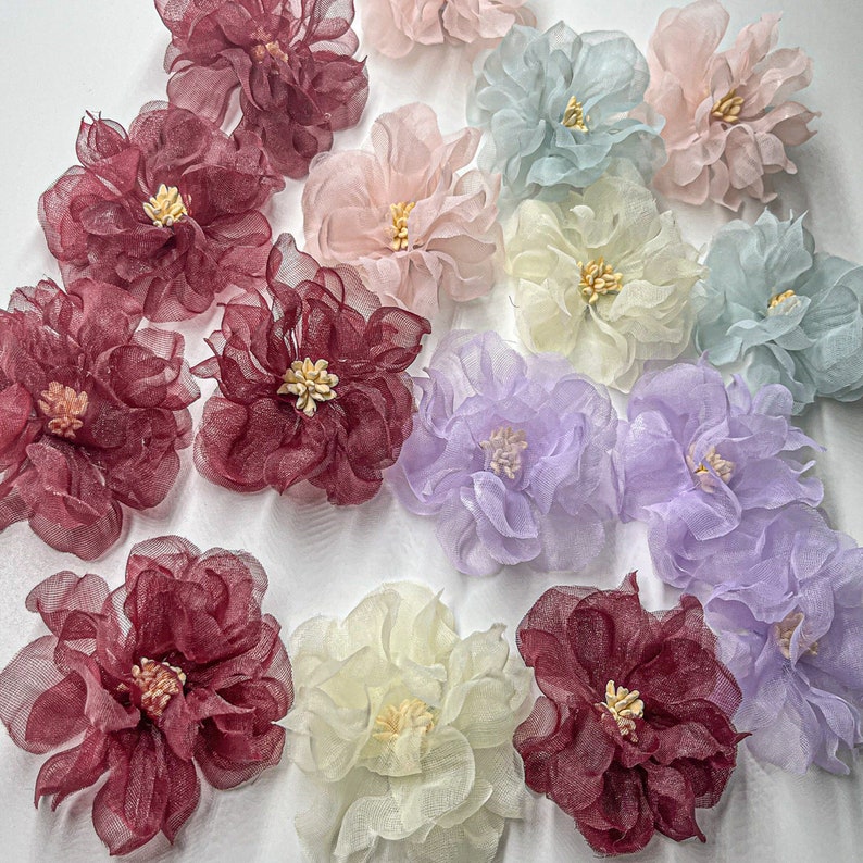 May include: Assortment of 12 delicate fabric flowers in various colors, including burgundy, lavender, cream, and light blue. The flowers are made of a soft, sheer fabric and have a layered, ruffled appearance.