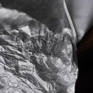 May include: Close-up of a crumpled, silver-colored plastic sheet. The plastic is wrinkled and has a metallic sheen.