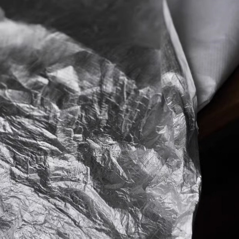 May include: Close-up of a crumpled, silver-colored plastic sheet. The plastic is wrinkled and has a metallic sheen.