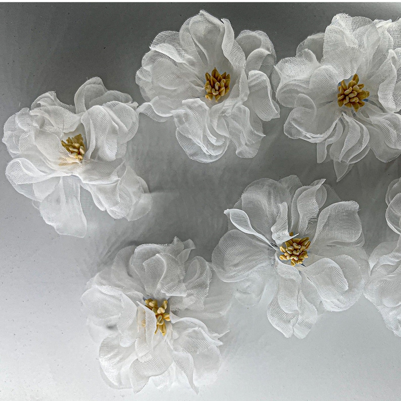 May include: Five white fabric flowers with gold centers. The flowers are arranged in a semi-circle on a gray background.