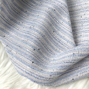 K&ouml;nnte beinhalten: Nahaufnahme eines hellblauen Stoffes mit einem dezenten wei&szlig;en und silbernen Streifenmuster. Der Stoff hat eine strukturierte, gewebte Optik und ist mit kleinen silbernen Pailletten verziert.
