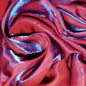 May include: Close-up of a red fabric with a subtle iridescent sheen. The fabric is folded and draped, creating soft, flowing lines.