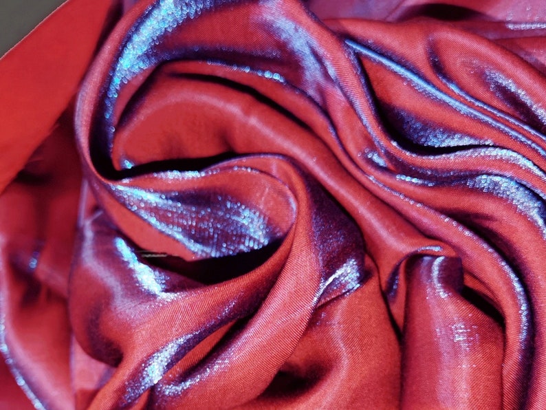 May include: Close-up of a red fabric with a subtle iridescent sheen. The fabric is folded and draped, creating soft, flowing lines.