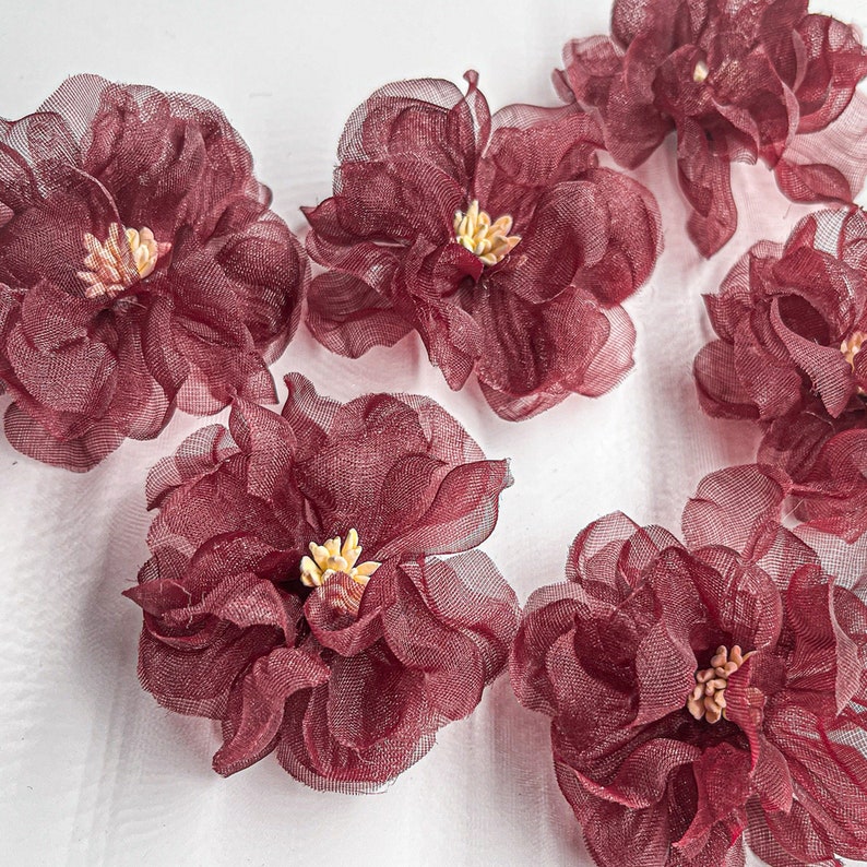 May include: Six burgundy-colored fabric flowers with a ruffled texture. The flowers have a light yellow center.